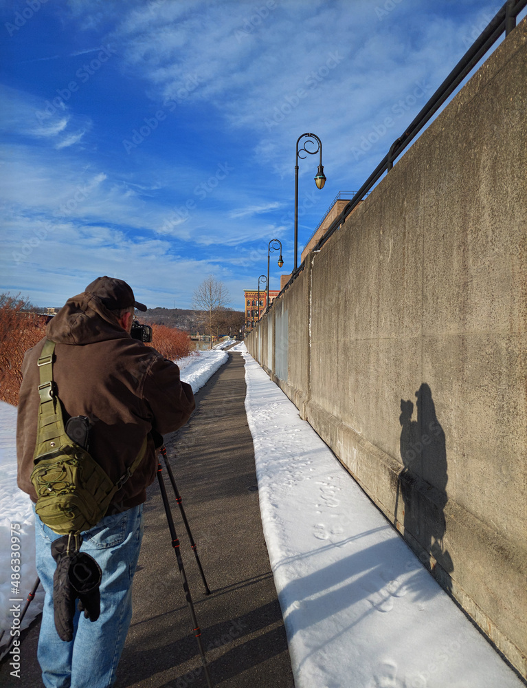 A male photographer with his camera and tripod takes photos while walking the Chenango River Walk in Binghamton in Upstate NY. A male photographer with his camera and tripod takes photos while walking the Chenango River Walk in Binghamton in Upstate NY.