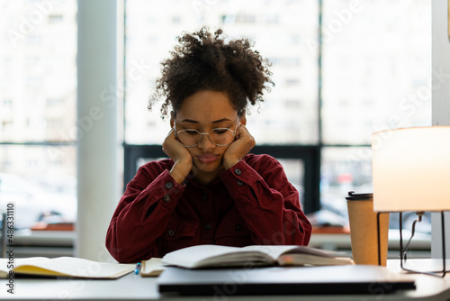 Waist up portrait view of the tired young woman feeling sleepy while reading a book and writing essay sitting at the table at the library