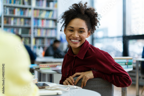 Waist up portrait view of the multiracial woman looking at the camera with toothy smile while spending time at the school library