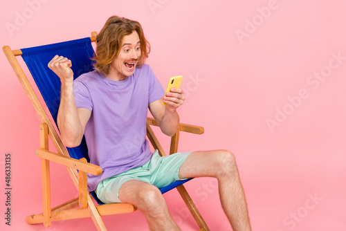 Portrait of handsome trendy cheery guy sitting in chair using device celebrating post smm like isolated over pink pastel color background