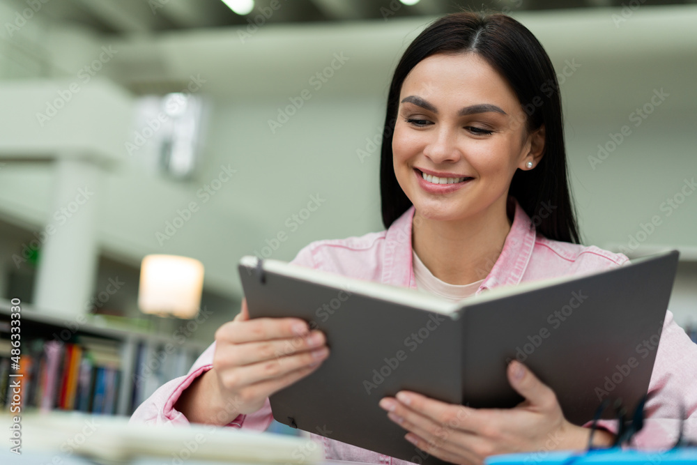 Low angle view of the brunette female student sitting in the library and reading book. High school student in the school library concept Low angle view of the brunette female student sitting in the library and reading book. High school student in the school library concept