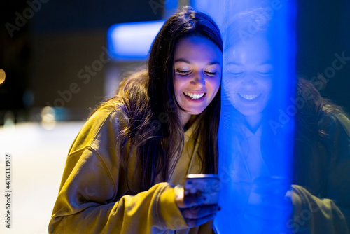  Friendly young woman with long hair next to a bluish neon wall looking at her mobile phone dressed in a yellow hoodie