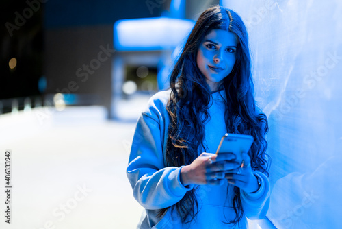Attractive and friendly young woman with long hair next to a bluish neon wall looking at her mobile phone dressed in a hoodie