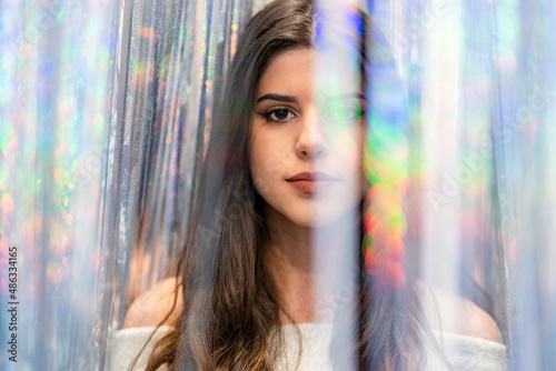 Portrait of a young woman with long hair looking at the camera between a curtain of silver and colored glitter and reflections