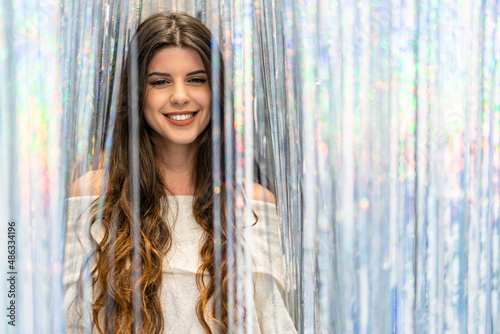 Portrait of a young woman with long hair looking at the camera between a curtain of silver and colored glitter and reflections