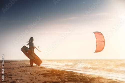 Kiteboarding. Kitesurfing athlete woman at sunset stands on the sandy shore holding her kite in the air and looks at the sea with waves and sunset