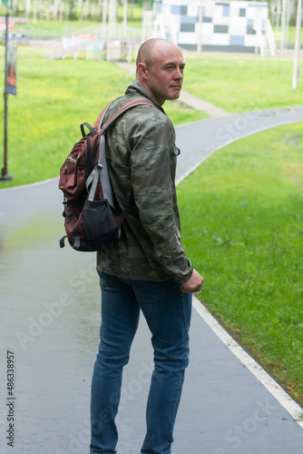 A sporty man in a military jacket and with a backpack in the park. Vertical photo.
