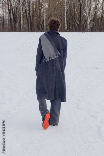 A young man in a long gray coat and scarf walks through the snow. Rear view. Back view.