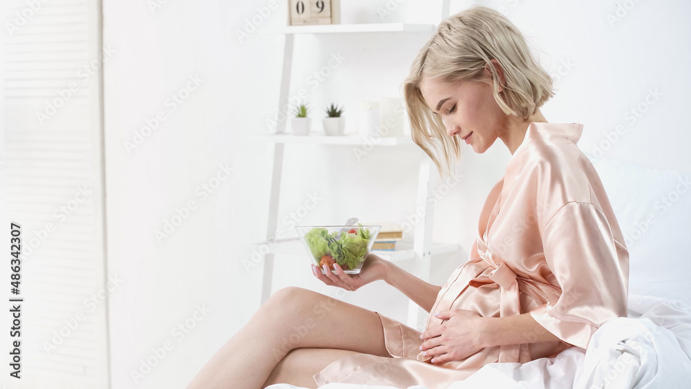 side view of pleased pregnant woman holding bowl with salad and looking at belly in bedroom side view of pleased pregnant woman holding bowl with salad and looking at belly in bedroom