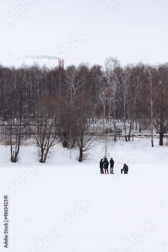 A small group of people on the frozen lake during a winter. One is sitting. Lots of trees without leaves and a smoke comming from the pipe far away in the background