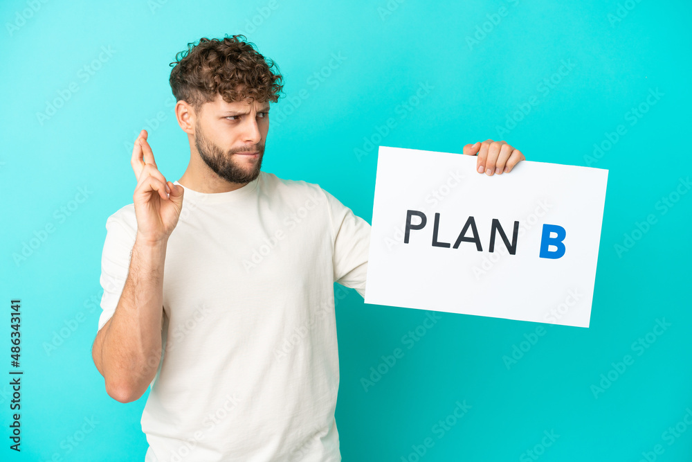 Young handsome caucasian man isolated on blue background holding a placard with the message PLAN B with fingers crossing Young handsome caucasian man isolated on blue background holding a placard with the message PLAN B with fingers crossing