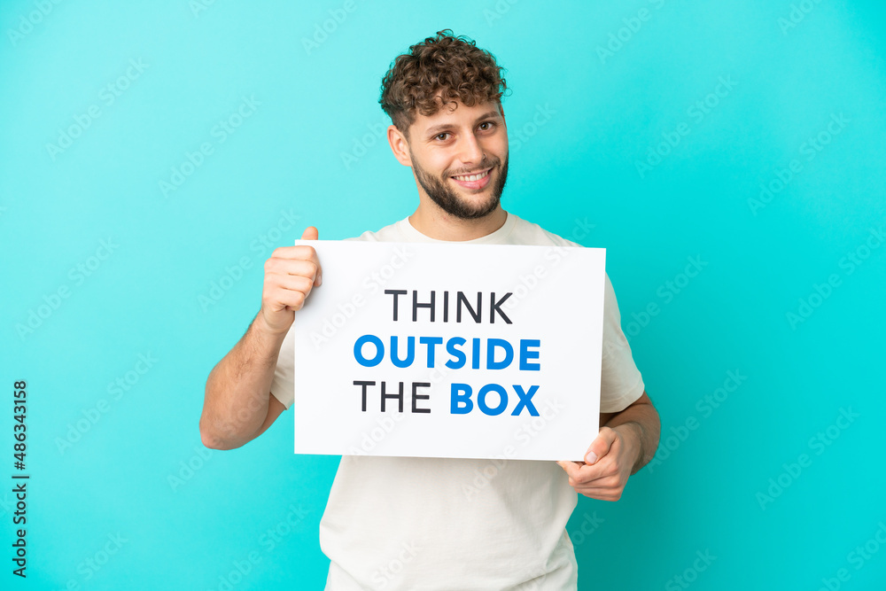 Young handsome caucasian man isolated on blue background holding a placard with text Think Outside The Box Young handsome caucasian man isolated on blue background holding a placard with text Think Outside The Box