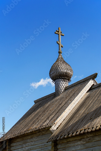 The wooden roof of the Orthodox chapel with a small wooden dome and a cross on it. Carved. Gray tree. Folk architecture. Clear blue sky. Daylight.
