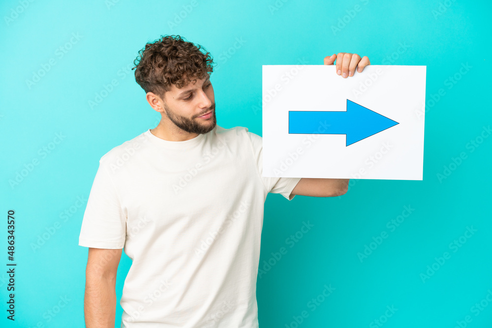 Young handsome caucasian man isolated on blue background holding a placard with arrow symbol Young handsome caucasian man isolated on blue background holding a placard with arrow symbol