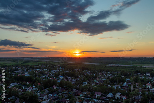 A view from a height of a large village on the background of a sunset with clouds. A motorway is visible in the distance. High-rise buildings are visible in the distance. Lots of clouds. Summer.