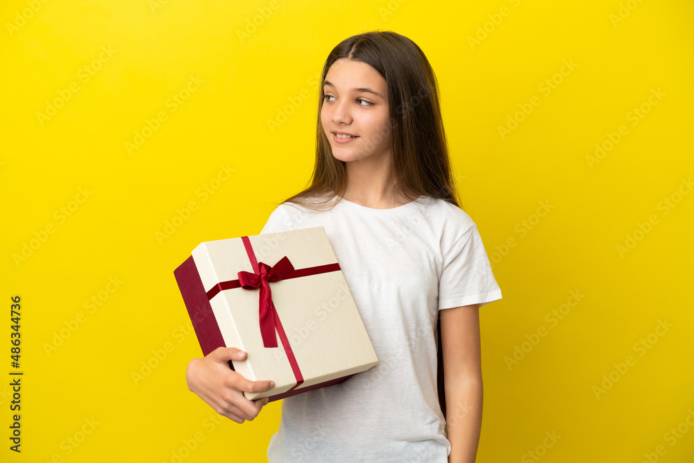 Little girl holding a gift over isolated yellow background looking to the side and smiling Little girl holding a gift over isolated yellow background looking to the side and smiling