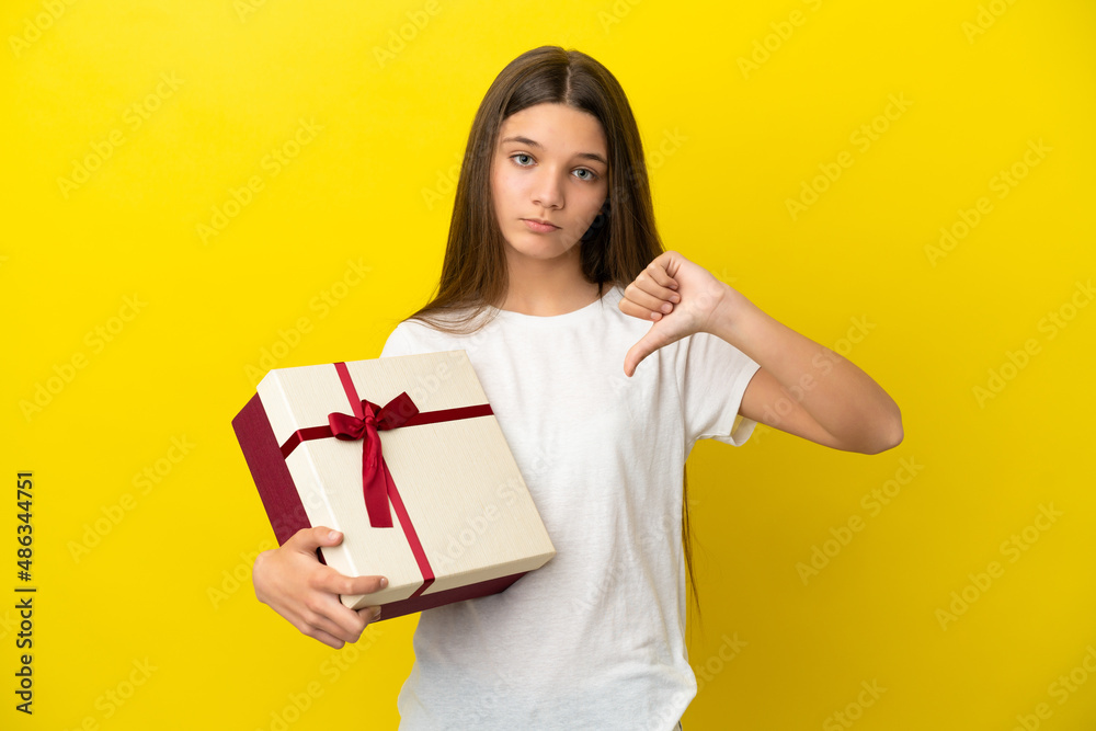 Little girl holding a gift over isolated yellow background showing thumb down with negative expression Little girl holding a gift over isolated yellow background showing thumb down with negative expression