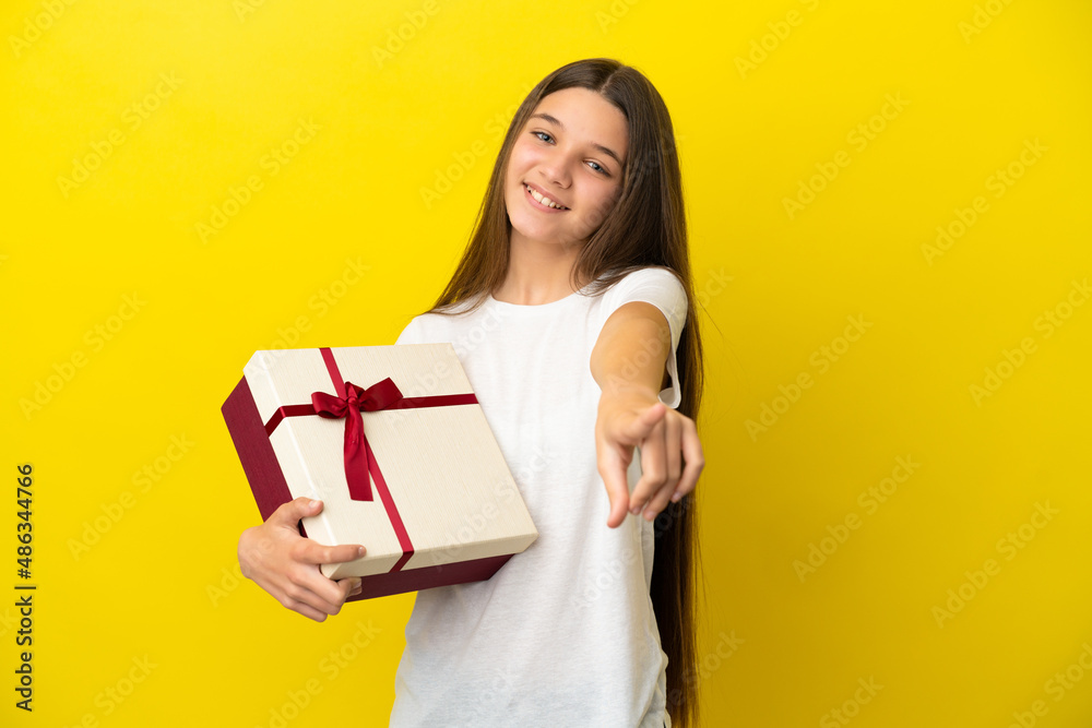 Little girl holding a gift over isolated yellow background pointing front with happy expression Little girl holding a gift over isolated yellow background pointing front with happy expression