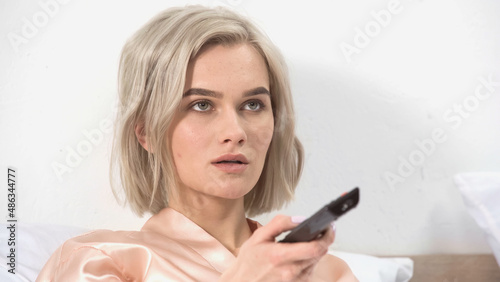 shocked blonde woman holding remote controller in bedroom