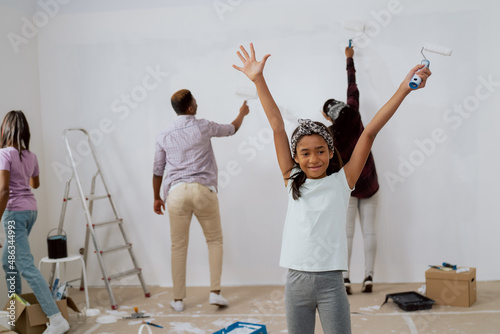 Little sweet adorable girl is happy to move to new apartment, helps in renovation of house, raises hands in the air with happiness, in the background parents and sister are painting walls of the room
