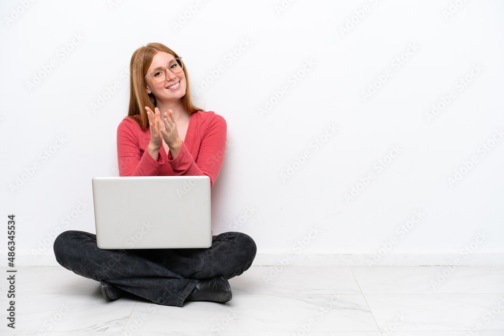 Young redhead woman with a laptop sitting on the floor isolated on white background applauding after presentation in a conference Young redhead woman with a laptop sitting on the floor isolated on white background applauding after presentation in a conference