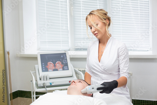 Photo of a young beautiful cosmetologist conducting a laser facial hair removal procedure with a modern device to a man lying on a couch in a cosmetology office