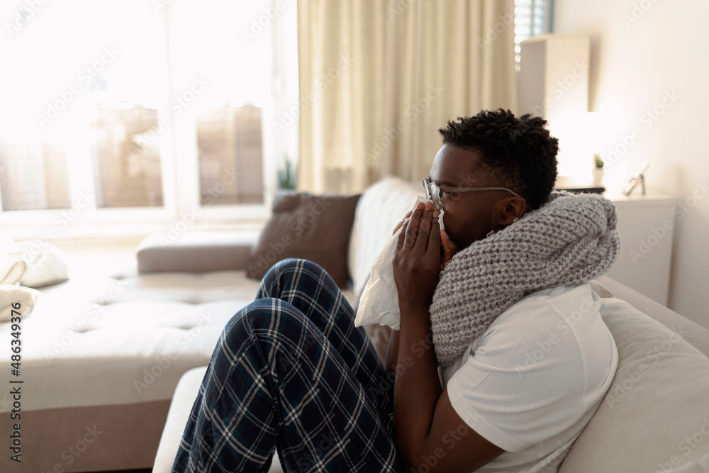 Young man sitting in his bed and blowing his nose while suffering from a cold. Sick African with cold or flu virus is sitting under the blanket on the sofa and blowing his nose with paper tissues. Young man sitting in his bed and blowing his nose while suffering from a cold. Sick African with cold or flu virus is sitting under the blanket on the sofa and blowing his nose with paper tissues.