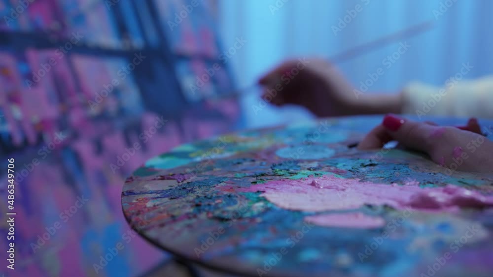 Focus on palette with oil paints. Artist draws picture on canvas in creative workshop. Woman dips brush into bright acrylic paints on palette and applies strokes to painting. Slow motion. Close up.