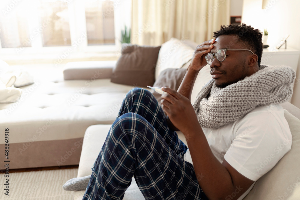 Fototapeta premium Man having a fever, measuring temperature with a digital thermometer. Man holding thermometer, measuring body temperature while suffering from influenza. Frustrated male with thermometer feeling sick.
