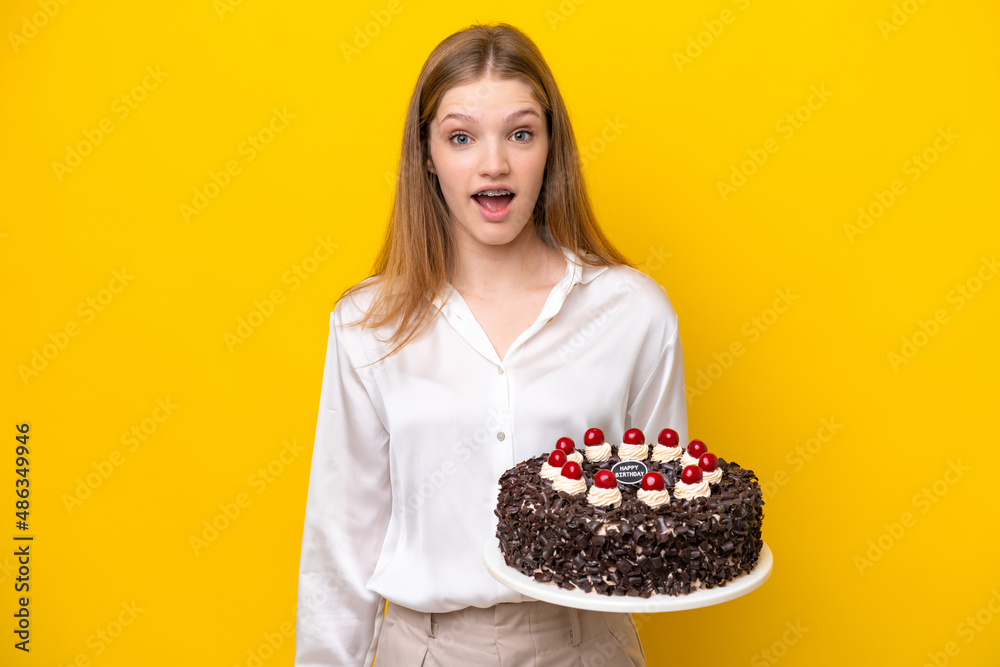 Teenager Russian girl holding birthday cake isolated on yellow background with surprise facial expression Teenager Russian girl holding birthday cake isolated on yellow background with surprise facial expression