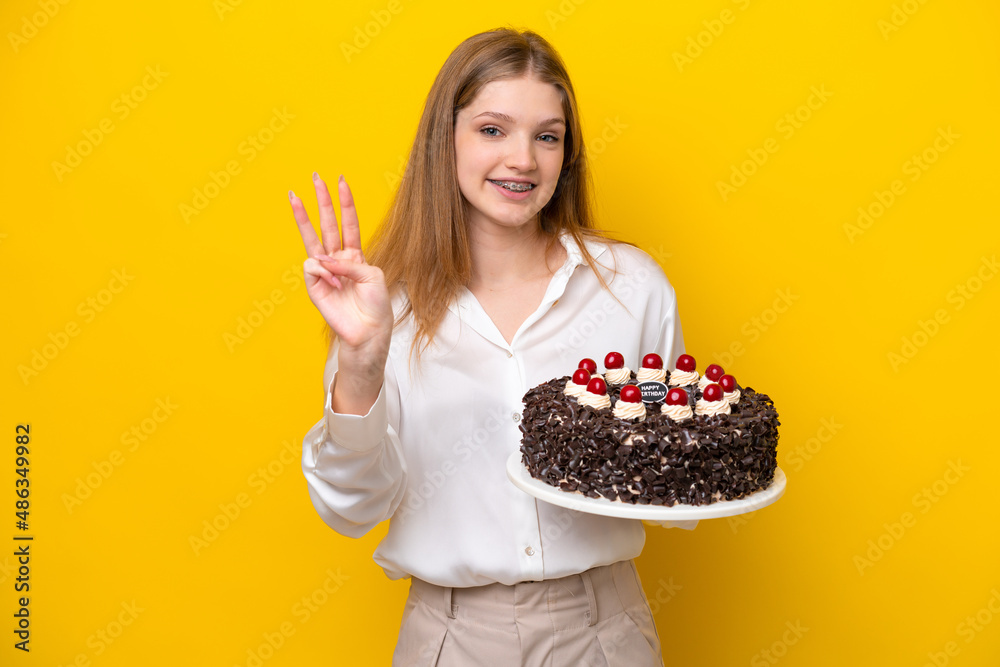 Teenager Russian girl holding birthday cake isolated on yellow background happy and counting three with fingers