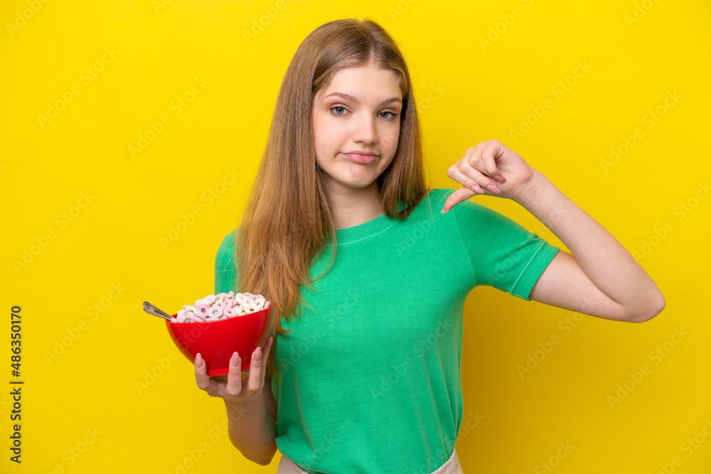 Teenager Russian girl holding bowl of cereals isolated on yellow background showing thumb down with negative expression Teenager Russian girl holding bowl of cereals isolated on yellow background showing thumb down with negative expression