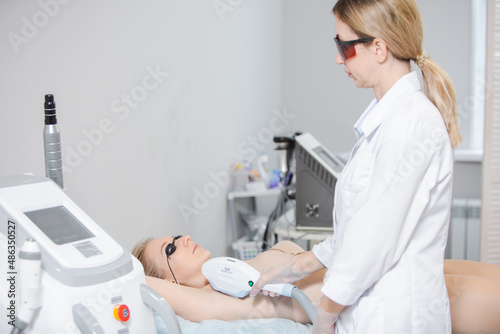 Photo of a modern cosmetology device and a female dermatologist in protective glasses performing a procedure of laser epilation of the armpits of a young girl