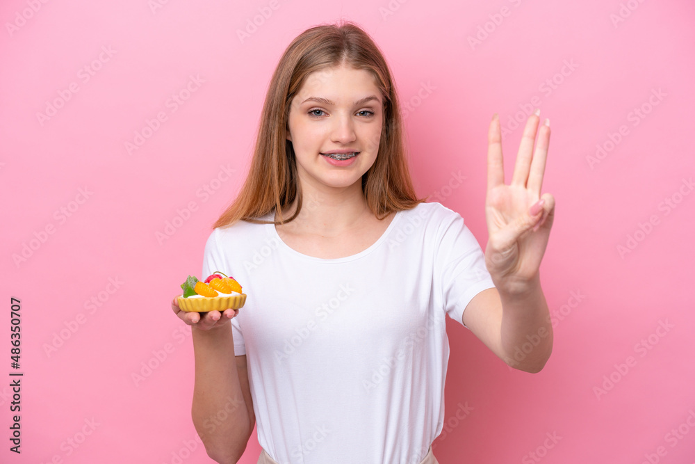 Teenager Russian girl holding a tartlet isolated on pink background happy and counting three with fingers Teenager Russian girl holding a tartlet isolated on pink background happy and counting three with fingers