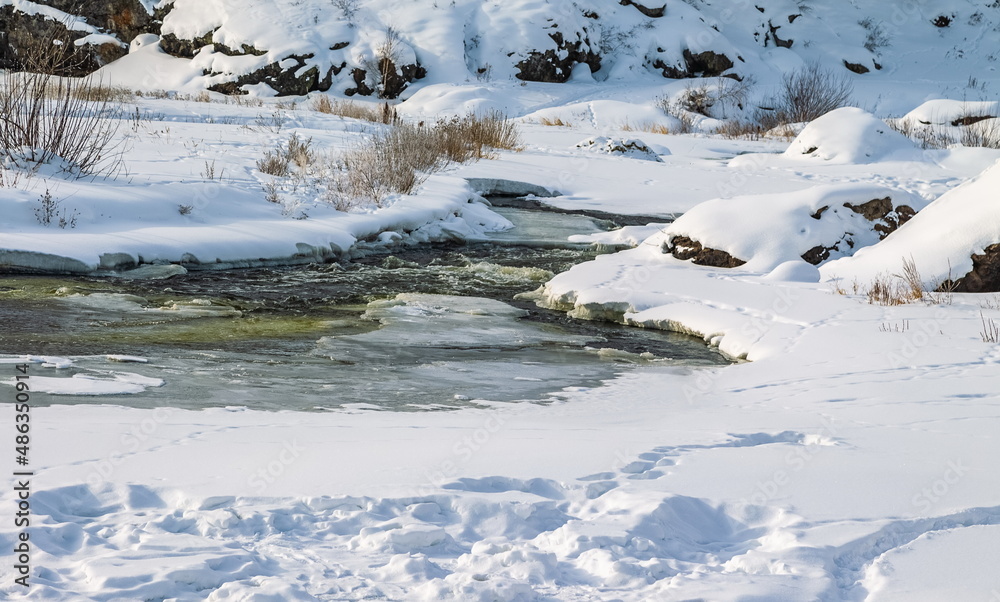 Winter landscape with a fast river with ice-free water, snow, dry grass and rocky shores Winter landscape with a fast river with ice-free water, snow, dry grass and rocky shores