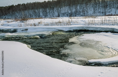 Winter landscape with a fast river with ice-free water, snow, dry grass and trees