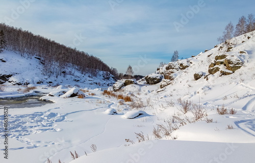 Winter landscape with a fast river with ice-free water, snow, dry grass and trees against a blue sky background