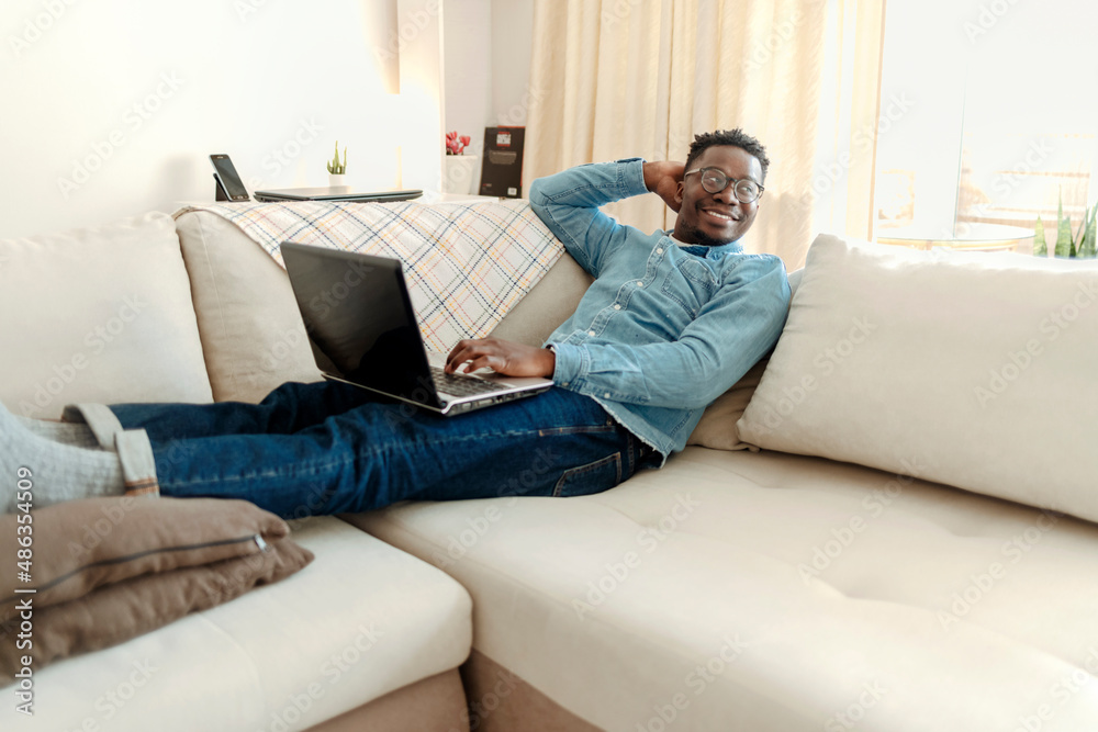 Handsome young African man looking at laptop and smiling while lying on the sofa at home. Shot of a handsome young man using a laptop at home.