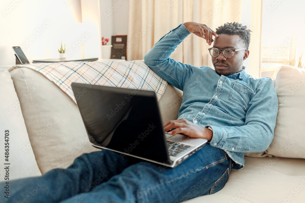 Handsome African man looking at laptop with serious face while lying on the sofa at home. One man, businessman working from home do to coronavirus pandemic outbreak, having online meeting on laptop. Handsome African man looking at laptop with serious face while lying on the sofa at home. One man, businessman working from home do to coronavirus pandemic outbreak, having online meeting on laptop.