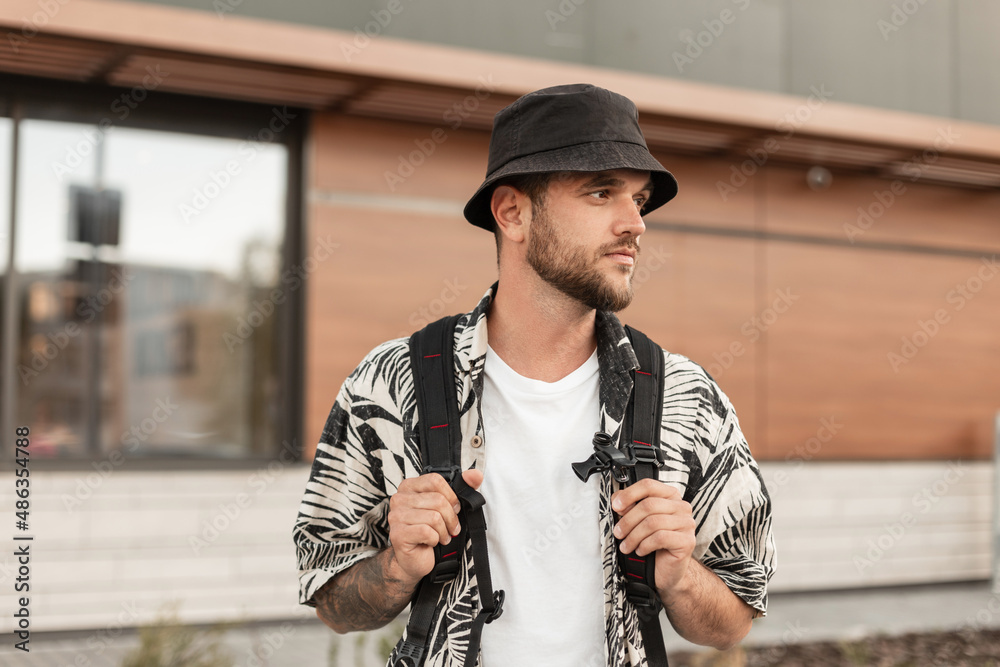 Stylish handsome hipster man with a bucket hat in fashionable clothes and a  shirt with a backpack walks in the city Stock Photo | Adobe Stock
