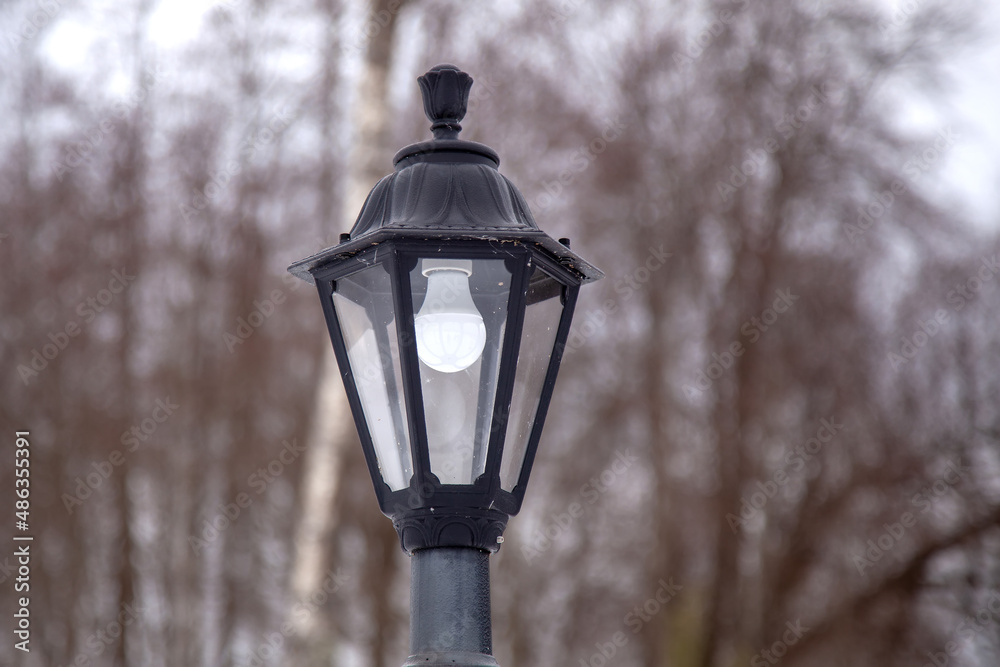A large iron lantern on a light blurry background close-up. Street lighting in dark areas of the city in winter. A light system in an open area. Forged large lamp in the yard.