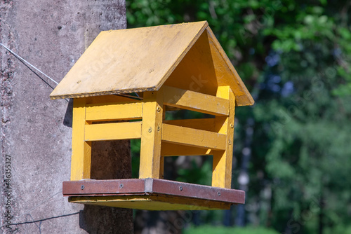 In summer, a yellow wooden bird feeder hangs on a tree. Creating places for self-feeding, where homeless and forest animals could eat. An improvised house in a city park. Birdhouse hand made