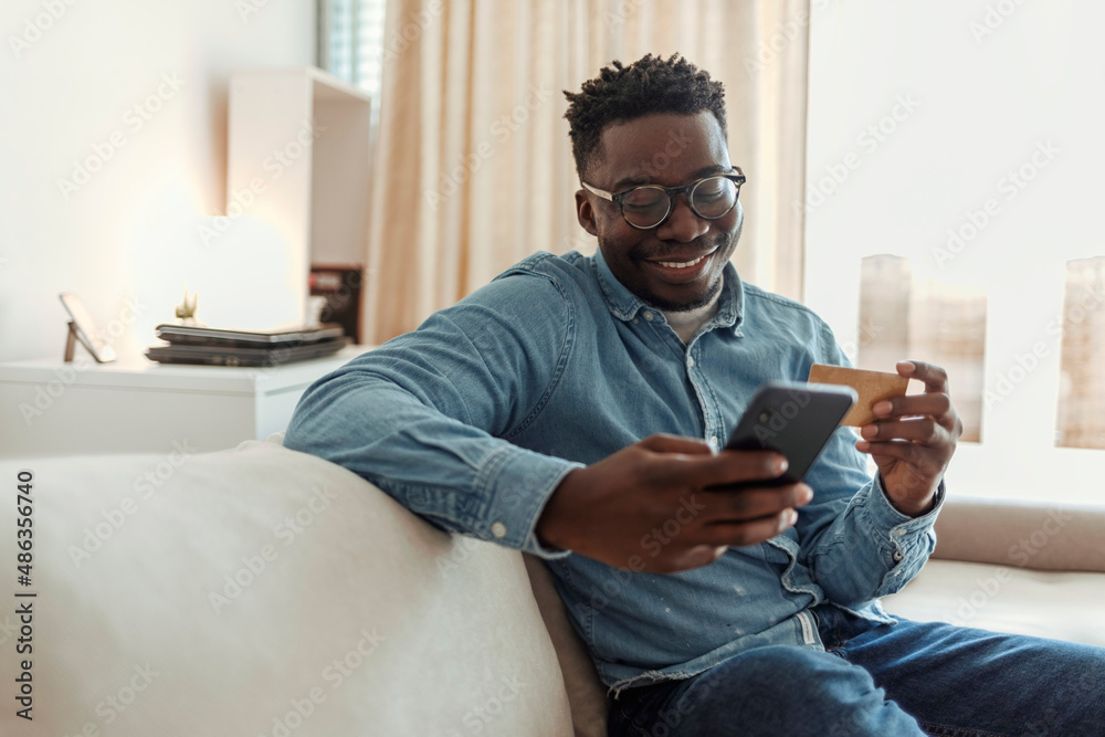 Shot of a young man holding his credit card while using a smartphone at home. Cheerful young man doing online shopping while using his cellphone and credit card inside of an apartment during the day.