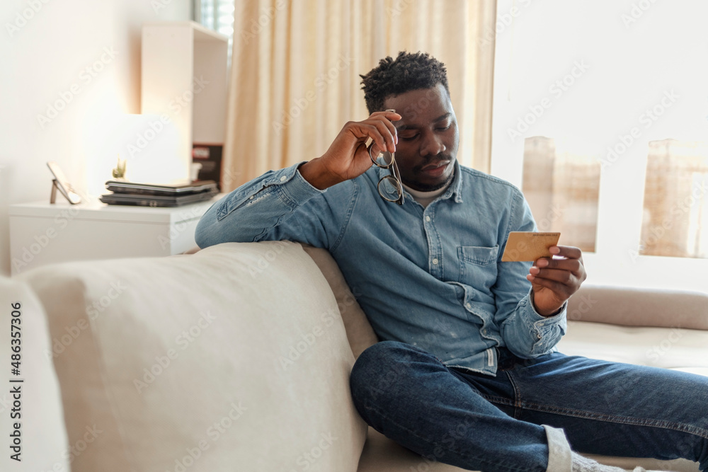 Confused young man having problem with blocked credit card making rejected unsecured online payment using phone at home. Upset men sitting on the couch, having problems with payment by credit card.