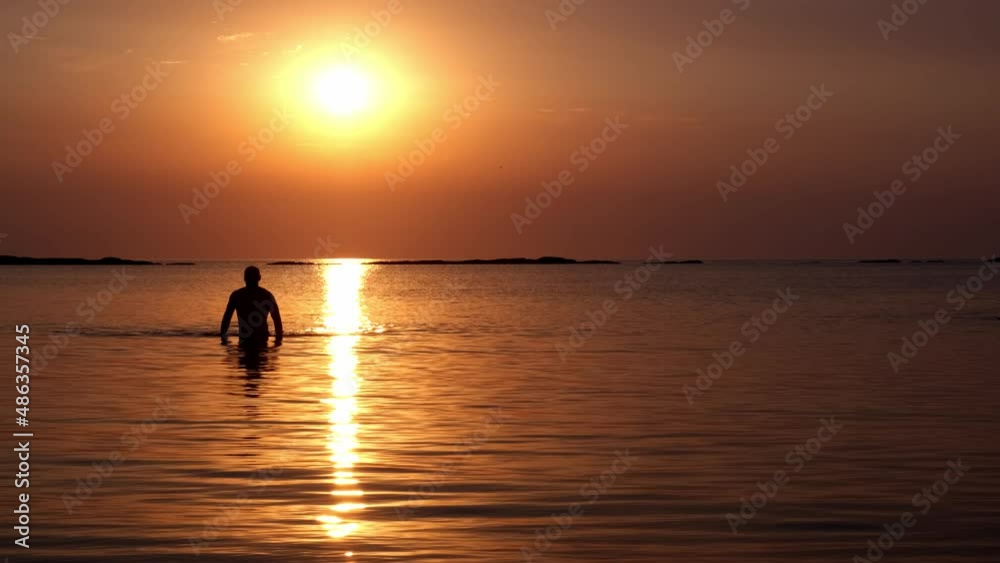 The silhouette of an athletic man comes out of the water and goes in the direction of the camera. The solar disk rises above the sea The silhouette of an athletic man comes out of the water and goes in the direction of the camera. The solar disk rises above the sea