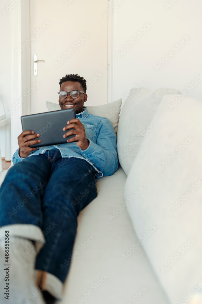 Shot of a handsome young man using his digital tablet while sitting on a sofa at home. Working with mobile gadget. Young African-American man using digital tablet, sitting on sofa at home, copy space Shot of a handsome young man using his digital tablet while sitting on a sofa at home. Working with mobile gadget. Young African-American man using digital tablet, sitting on sofa at home, copy space