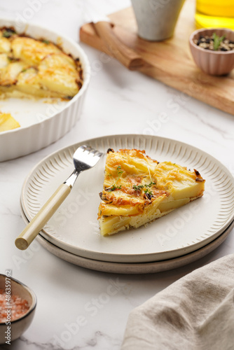 Slice of potato casserole with red onions, fresh thyme and rosemary on green checkered kitchen towel , wooden board with flavoring ingredients: pepper, salt, olive oil