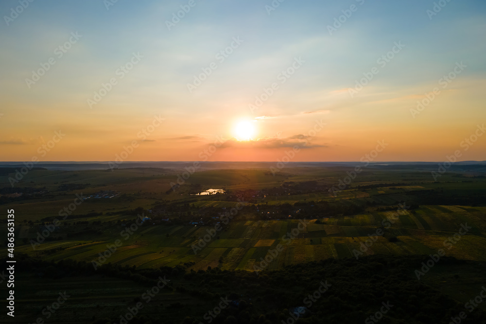 Aerial landscape view of green cultivated agricultural fields with growing crops and distant village houses on bright summer evening