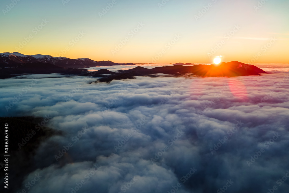 Aerial view of vibrant sunrise over white dense clouds with distant dark mountains on horizon