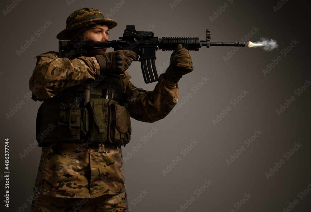 Portrait of soldier in military camouflage uniform protected with helmet, body armor, holding machine gun desaturated on a gray background. Army. Sniper. Soldier shooting. Military Conflict Actions. Portrait of soldier in military camouflage uniform protected with helmet, body armor, holding machine gun desaturated on a gray background. Army. Sniper. Soldier shooting. Military Conflict Actions.